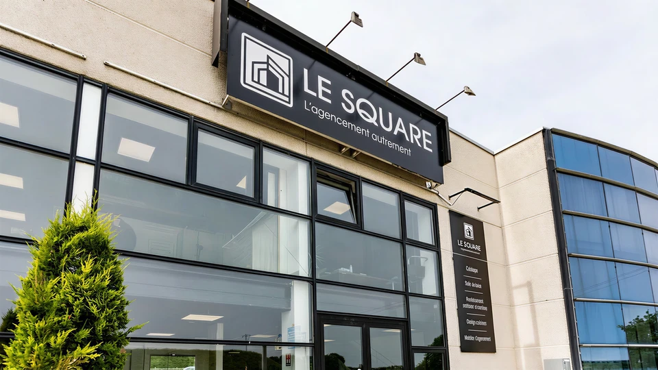 Intérieur moderne d'un showroom de luxe : espace lumineux et épuré de 500 m² avec exposition de cuisines haut de gamme, dressings sur mesure, carrelages italiens et espagnols, revêtements de sol premium et meubles de salle de bain design. L'ambiance est claire et professionnelle, avec des matériaux de qualité visibles, une mise en scène soignée, et un détail subtil montrant deux personnes en conversation (client et spécialiste) qui explore les matériaux côte à côte, illustrant le conseil personnalisé dans un environnement privatisé et tranquille.